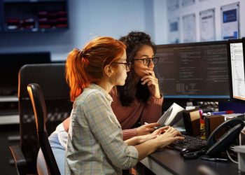 Two developers collaborate on a project as they review code on a display in their workspace.